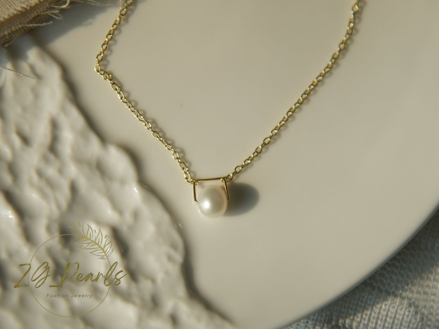 Gold necklace with a pearl pendant on a textured surface, featuring the brand 'ZG Pearls'.