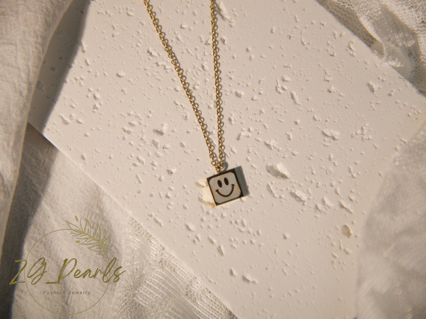 Gold necklace with a smiley face pendant on a textured beige surface, featuring the brand '24 Pearls'.