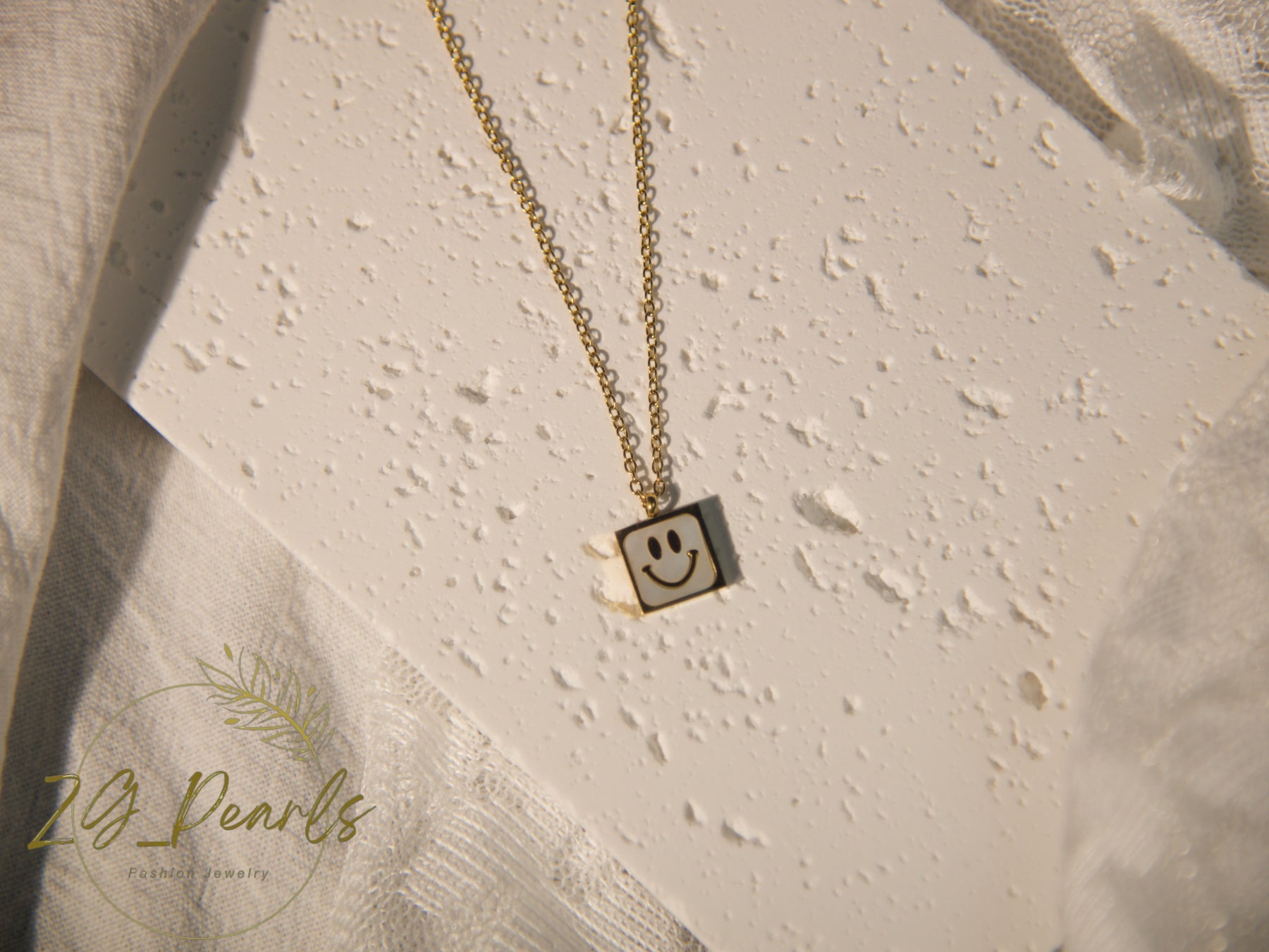 Gold necklace with a smiley face pendant on a textured beige surface, featuring the brand '24 Pearls'.