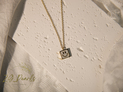 Gold necklace with a smiley face pendant on a textured beige surface, featuring the brand '24 Pearls'.