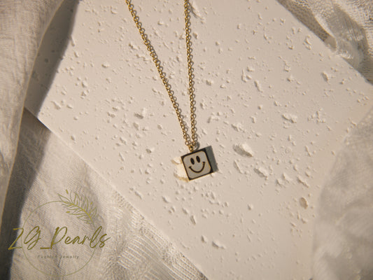 Gold necklace with a smiley face pendant on a textured beige surface, featuring the brand '24 Pearls'.