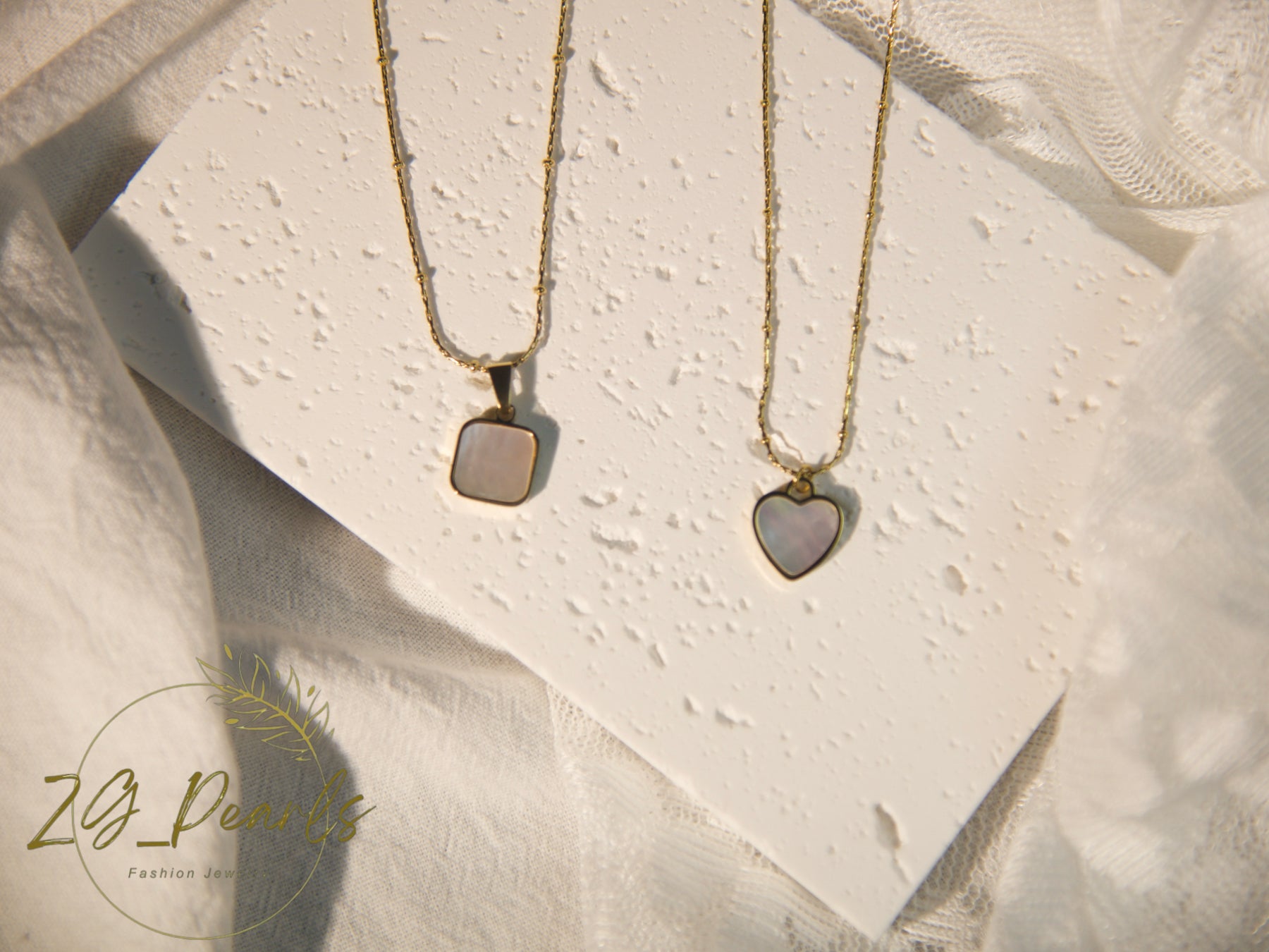 Two gold necklaces with heart and square pendants on a textured white surface.